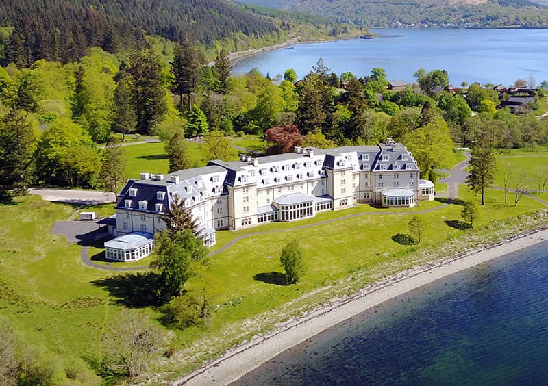 Aerial view of the Ardgartan Hotel bathed in bright sunshine, surrounded by lush greenery and overlooking the serene waters of Loch Long