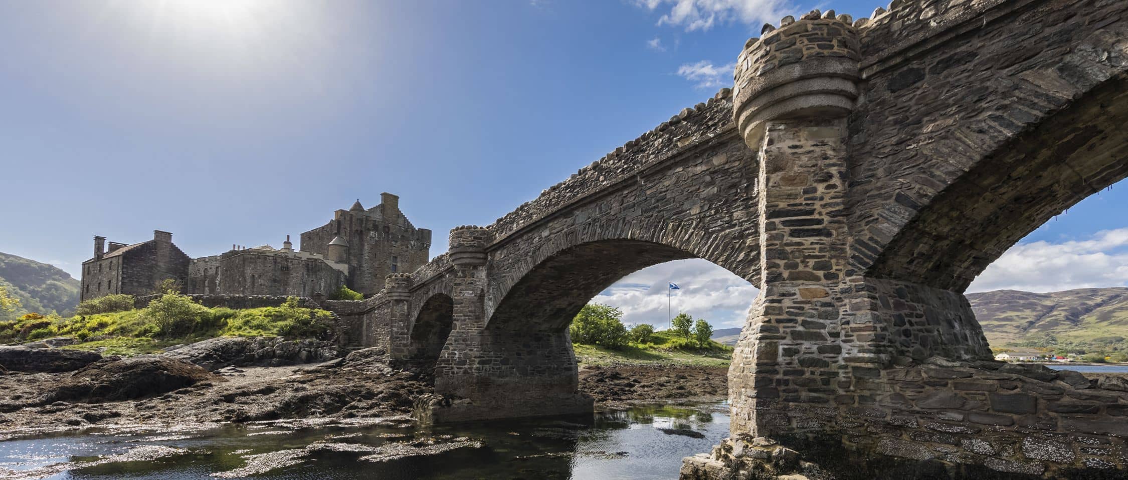 A stone bridge leads to Eilean Donan Castle, an iconic Scottish landmark set against a backdrop of sunshine and reflective waters.